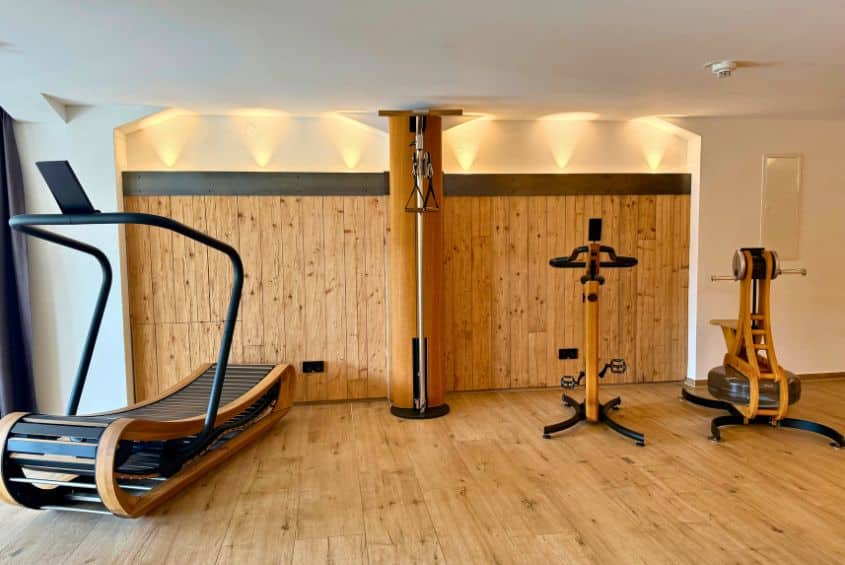 Frontale Ansicht des Fitnessbereichs mit hochwertigen, motorlosen Geräten der Marke NOHRD aus hellem Holz, darunter ein Laufband links, ein Wand-Kabelzug in der Mitte und ein Ergometer sowie WaterGrinder rechts.
