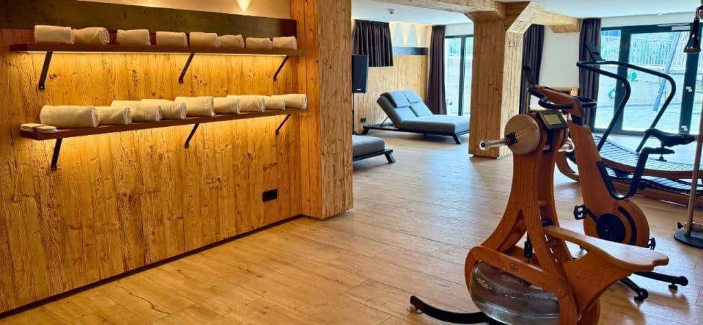 Blick in den hellen, holzverkleideten Wellnessbereich mit einem Regal voller frischer, weißer Handtücher links, ergonomischen grauen Ruheliegen im Hintergrund und NOHRD-Fitnessgeräten aus Holz im Vordergrund.